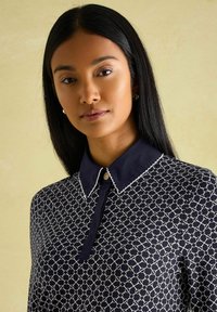 Marineblaue Bluse mit weißem geometrischem Muster, ausgestattet mit einem spitzen Kragen, Knopfverschluss und kontrastierendem Kragenbesatz. Glatter Stofftextur.