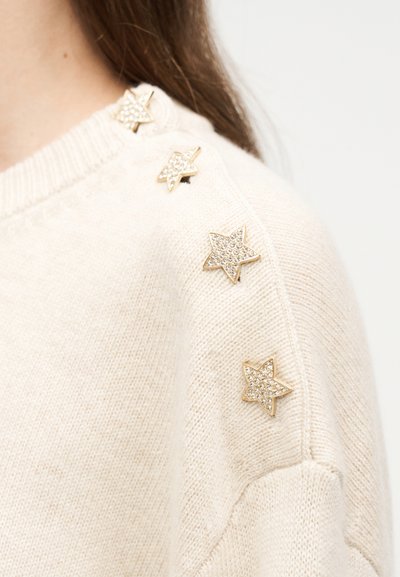 Κιτρινωπή πλεκτή μπλούζα με ώμο διακοσμημένο με τέσσερα χρυσούς αστεροειδείς στολίδια με μικρούς διαυγείς κρυστάλλους.