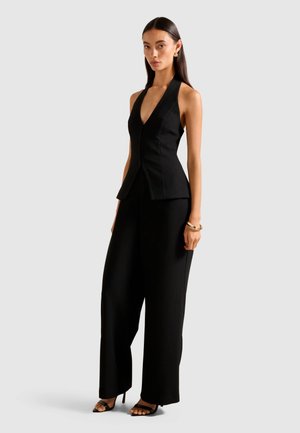 Woman wearing a sleeveless black halter top and wide-leg black trousers, accessorized with gold hoop earrings and bracelets, standing against gray.
