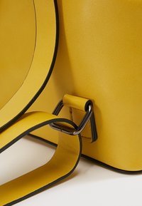 Sac en simili cuir jaune avec une texture lisse, présentant un bord accentué noir et une sangle fixée par une attache métallique argentée.