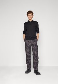 Camisa polo preta de manga longa, combinada com calças cargo cinzas estampadas que apresentam bolsos laterais e um corte relaxado. Botas pretas com solas texturizadas.