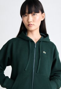 Grønn zip-up hettegenser laget av mykt stoff, med kengurulomme og snor. En liten logo er plassert på venstre brystområde.