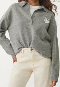 Sudadera gris de punto con cuello, tres botones y un diseño bordado en el pecho. Combinada con vaqueros de color beige claro.