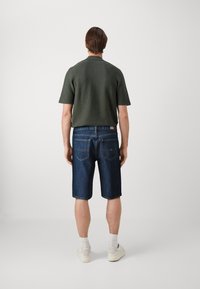 Polo de color caqui con mangas cortas combinado con pantalones cortos de mezclilla azules. Los pantalones cortos tienen dos bolsillos traseros y un corte recto. Zapatillas deportivas blancas y calcetines.