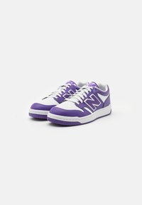 Fioletowe i białe trampki z perforowanym noskiem, sznurowanym wzorem i logo New Balance. Posiadają teksturowaną gumową podeszwę i kontrastowe akcenty.
