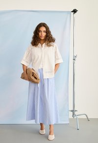 Jauna moteris su garbanotais plaukais, vilkinti baltą siuvinėtą palaidinę, mėlynos ir baltos spalvų dryžuotas plačias kelnes, rankose laiko rudą megztą delninę, stovi studijoje.