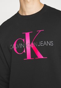 Sweatshirt noir en coton avec un grand texte brodé en rose et argent : "CK CALVIN KLEIN JEANS." Encolure ronde et manches longues.