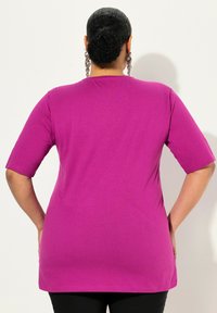 Maglietta magenta aderente con maniche corte, texture liscia e orlo dritto, che mostra la schiena con le mani nelle tasche.