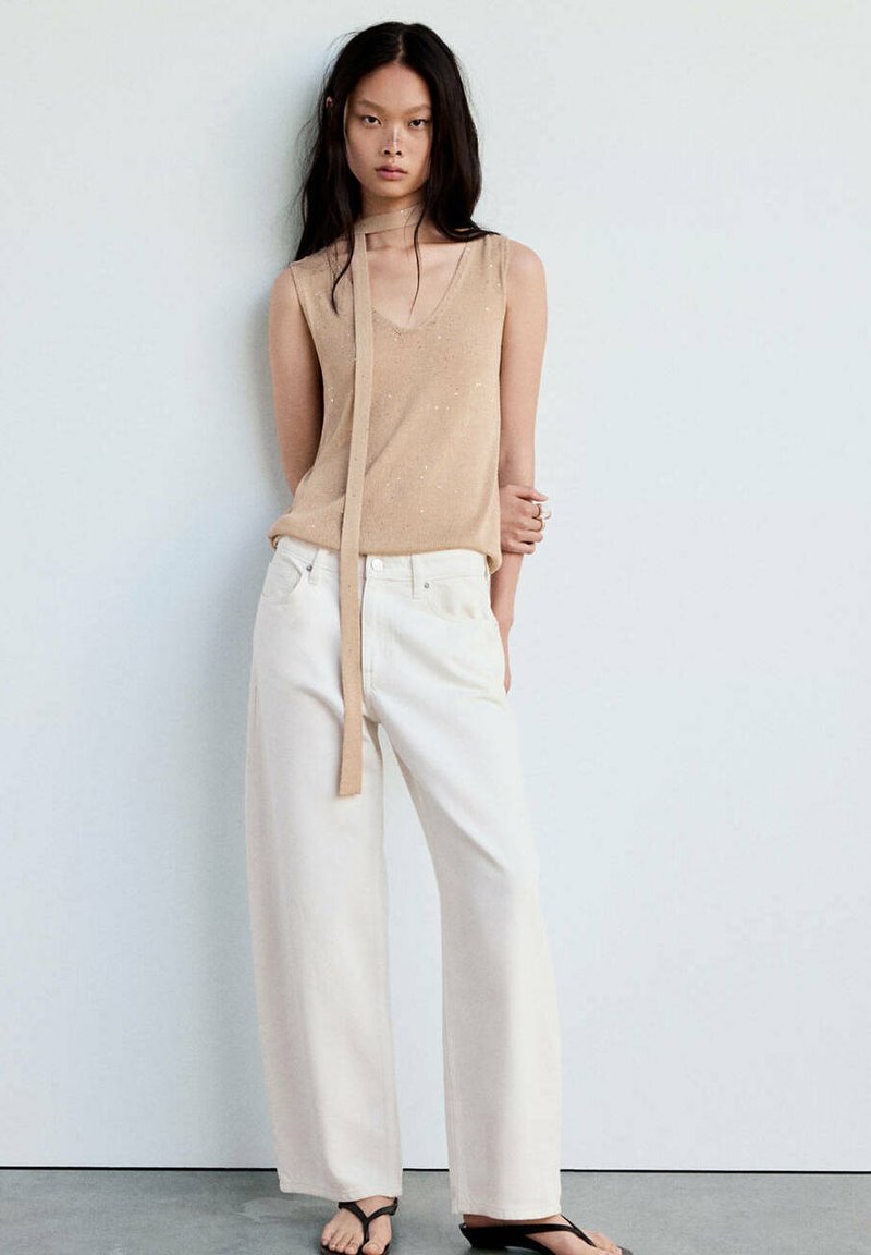 Femme debout avec les bras derrière le dos, portant un haut beige sans manches et un pantalon blanc à jambes larges sur un fond uni.