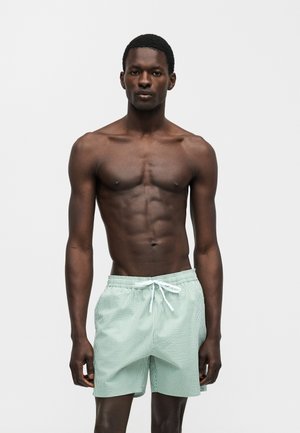 Άντρας με τονισμένους μυς που φοράει ανοιχτοπράσινα και λευκά ριγέ σορτς με κορδόνι, στέκεται μπροστά σε απλό φόντο.