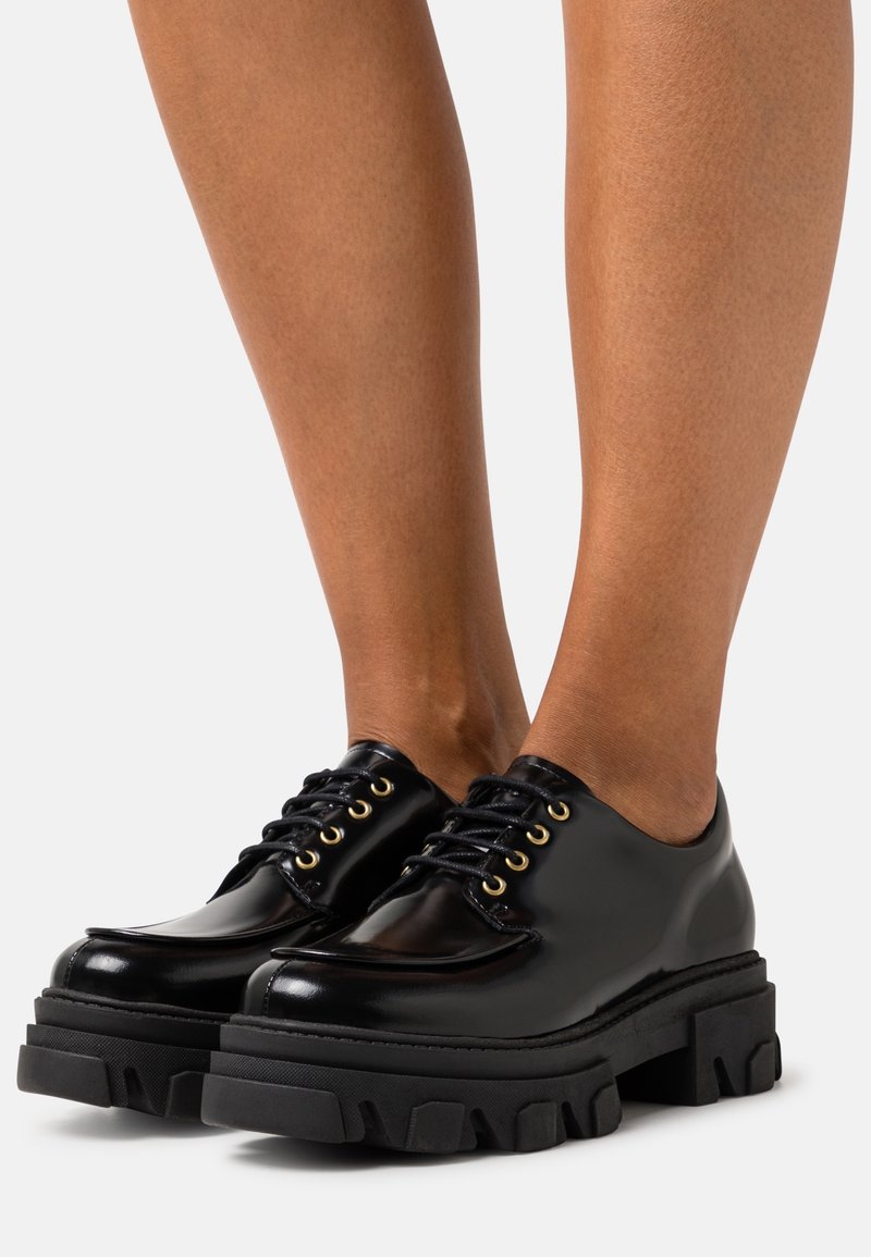 Chaussures à plateforme épaisses noires avec lacets portées sur des pieds à peau lisse, présentées sur un fond clair uni.