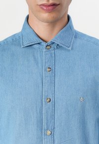 Camicia di jeans azzurro chiaro con trama testurizzata, dotata di colletto a punta, bottoni argentati e un piccolo logo ricamato sul petto.