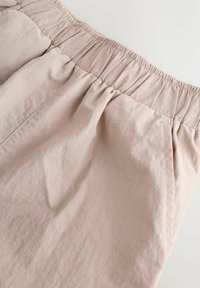 Šviesaus fono kreminės spalvos kelnės su elastinga juosmens dalimi, susmeigtais audinio raukiniais ir šoninėmis kišenėmis.