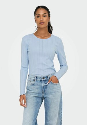 Joven mujer con una blusa de manga larga azul claro y jeans de cintura alta, de pie con confianza con una mano en la cadera y un fondo neutro.