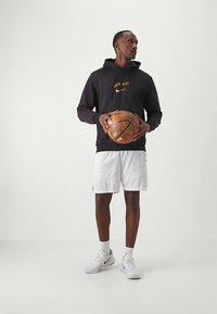 Schwarzer Hoodie mit gelbem Nike-Logo und Schriftzug, kombiniert mit weißen Shorts. Hält einen Basketball, trägt weiße Sneaker und Socken.