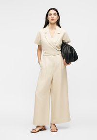 Γυναίκα με μακριά σκουρόχρωμα μαλλιά φοράει ένα μπεζ ολόσωμο κοντομάνικο ρούχο και σανδάλια, κρατώντας μια μεγάλη μαύρη δερμάτινη τσάντα.