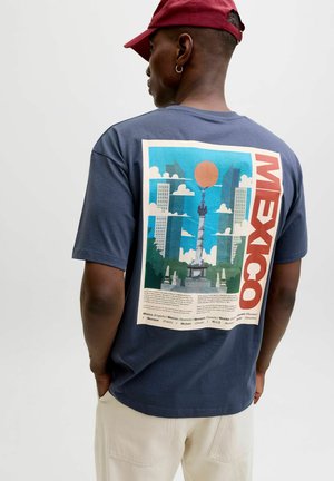 Homme portant une casquette rouge et un t-shirt bleu marine avec une scène colorée de la ville de Mexico et le mot "MEXIQUE" en grandes lettres au dos.