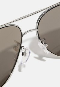 Óculos de sol aviador com lentes cinzentas escuras, armação de metal prateado e almofadas nasais transparentes. A marca Michael Kors é visível na armação.