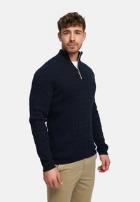 Marineblå kabelstrikket genser med høy krage og halv glidelås. Har teksturert stoff og ribbestrikkede mansjetter. Bruker khakibukser.