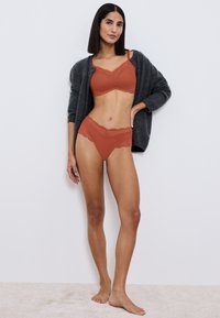 Terracotta kant bralette en bijpassende slipjes, beide met schulpachtige randen. Model draagt een donkergrijze cardigan over het set. Neutrale achtergrond.