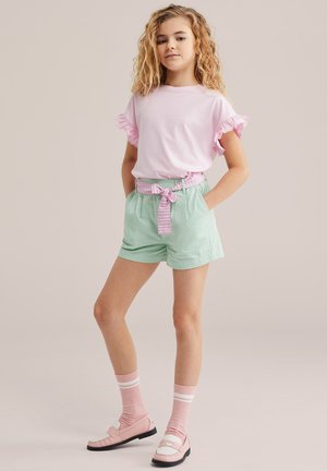 Fille debout les mains dans les poches portant une chemise rose à manches à volants, un short vert menthe avec une ceinture rayée, des chaussettes roses et des chaussures roses.