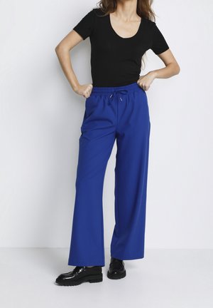 Femme portant un haut noir à manches courtes, un pantalon large bleu vif avec cordon de serrage, et des mocassins noirs, debout les mains sur les hanches.