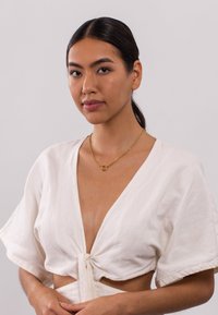 Haut blanc court à manches courtes, noué à l'avant. Porté avec un collier en chaîne dorée avec un pendentif circulaire. Texture lisse.