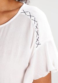 Blusa bianca con maniche corte e leggere. Presenta ricami neri a punto X sulla spalla. Tessuto morbido e leggero con una finitura testurizzata.