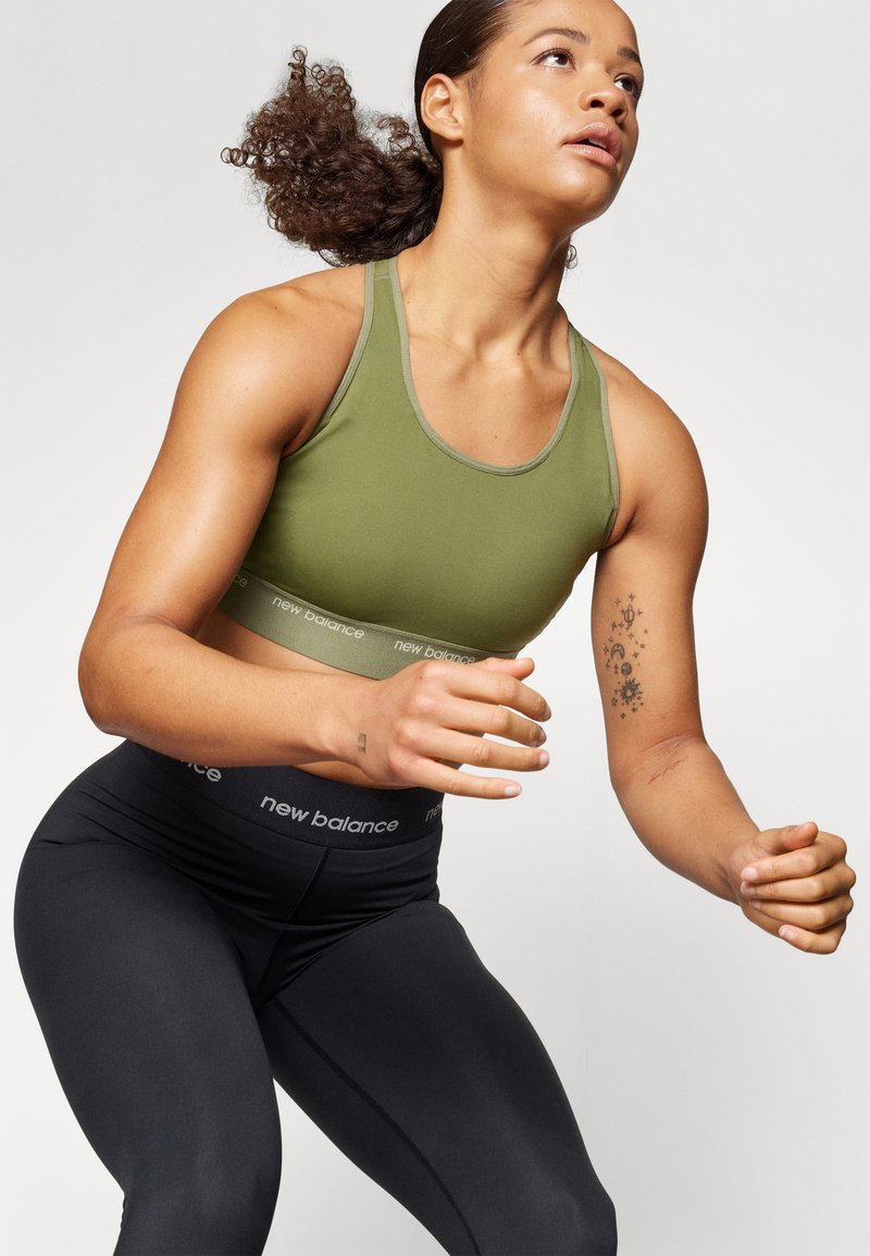 Athletische Frau in grünem Sport-BH und schwarzen Leggings mit New Balance-Branding, in der Bewegung vor einem schlichten weißen Hintergrund.