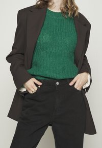 Persona che indossa un blazer marrone scuro sopra un maglione di lana verde, inserito in jeans neri a vita alta, con le mani in tasca e anelli d'oro.