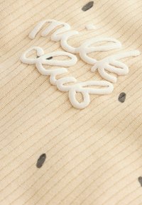 Krém színű bordás textilem, fehér hímzett szöveggel és fekete pöttyös részletekkel. A közelkép kiemeli a textúrát és a varrást.