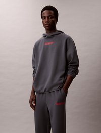 Szara bluza z kapturem z czerwonym logo "CALVIN KLEIN" na klatce piersiowej, połączona z pasującymi szarymi dresami z logo na biodrze. Gładka tekstura.