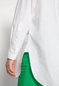 Gros plan d'une personne portant une chemise blanche à manches longues avec des poignets boutonnés et un pantalon vert, montrant le détail de la fente latérale de la chemise.