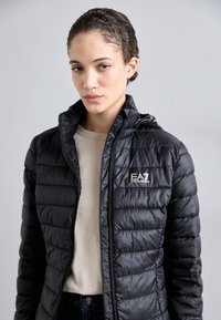 Črna puhovka z zadrgo, ki vključuje kapuco in vertikalno prešito zasnovo. Logotip "EA7 Emporio Armani" na levem prsnem delu.