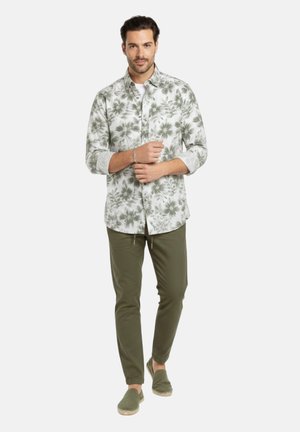 Hombre vistiendo una camisa de botones con estampado floral verde, pantalones verde oliva y zapatos verdes sin cordones, de pie con los brazos cruzados, mirando hacia adelante.