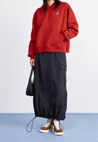 Sudadera roja con capucha y bolsillo frontal con logo blanco, combinada con una falda larga negra con bolsillos laterales y dobladillo elástico, complementada con zapatillas altas marrones.