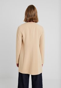 Abrigo beige a medida con mangas largas, textura suave y corte recto. Diseño minimalista, con un ligero detalle de costura en la parte trasera.