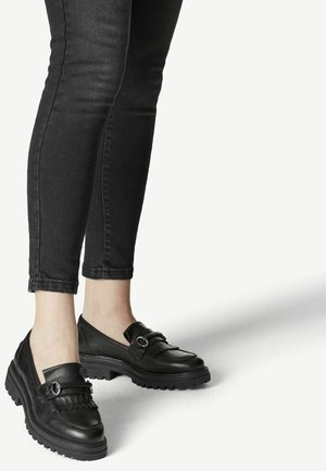 Ben iført tætsiddende sorte jeans og sorte læder loafers med chunky såler og sølvspænde på lys baggrund.