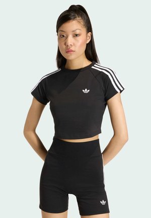 Joven mujer con coleta alta negra, vistiendo un crop top negro de Adidas y shorts de talle alto a juego con franjas blancas y logos, de pie con las manos detrás de la espalda.