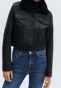 Femme portant une veste en cuir noire courte avec une fermeture à boutons et des poches, associée à un jean bleu.