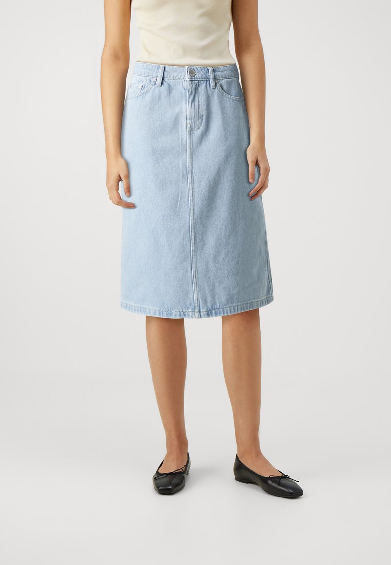 Világoskék denim midi szoknya egyenes szabással, elöl varrással és öt zsebbel. Fekete lapos cipővel és testre szabott krém színű toppal viselve.