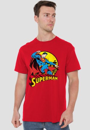 Young man wearing a red t-shirt with a vintage Superman graphic and yellow cityscape background, looking to his left.
