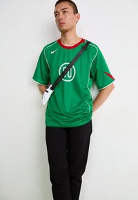 Groene sporttrui met rode accenten en een wit logo, gecombineerd met zwarte broeken en een kleine witte crossbody tas. De trui heeft een textuurpatroon.