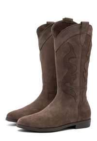 Botas de vaquero de ante marrón con un diseño curvado y costuras decorativas. La suela es plana con un ligero tacón. Tiene tiradores dobles en el caña.