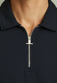 Tricou polo bleumarin dintr-un material texturat, cu închidere prin fermoar la jumătate și un trage fermoar din argint. Gulerul este plat și structurat.