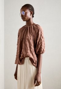 Braune texturierte Bluse mit gerafftem Ausschnitt, puffigen Ärmeln, kombiniert mit einem hellen cremfarbenen Rock. Goldene Sonnenbrille und schlichte Schmuckaccessoires.