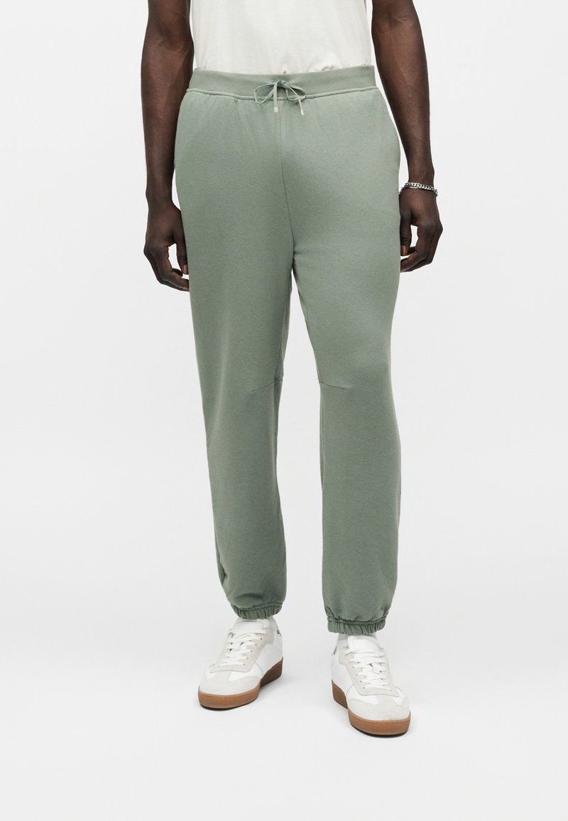 Homme portant un pantalon de jogging vert clair, des baskets blanches et une chemise blanche, debout devant un fond gris uni.