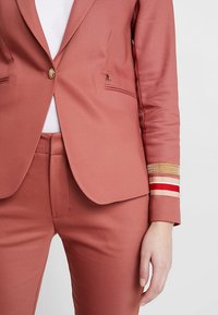 Rostfärgad kavaj med skräddarsydd design, utrustad med guldknapp, två framfickor och randig accent på ärmen i röd, beige och guld.