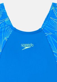 Maillots de bain bleus avec un devant bleu uni et des motifs végétaux complexes en bleu clair et jaune sur les côtés. Logo "Speedo" en jaune.