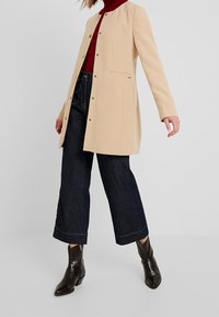 Abrigo de mezcla de lana beige con cuello alto, botones de presión y bolsillo frontal. Combinado con vaqueros azul oscuro de pierna ancha y botines marrones.